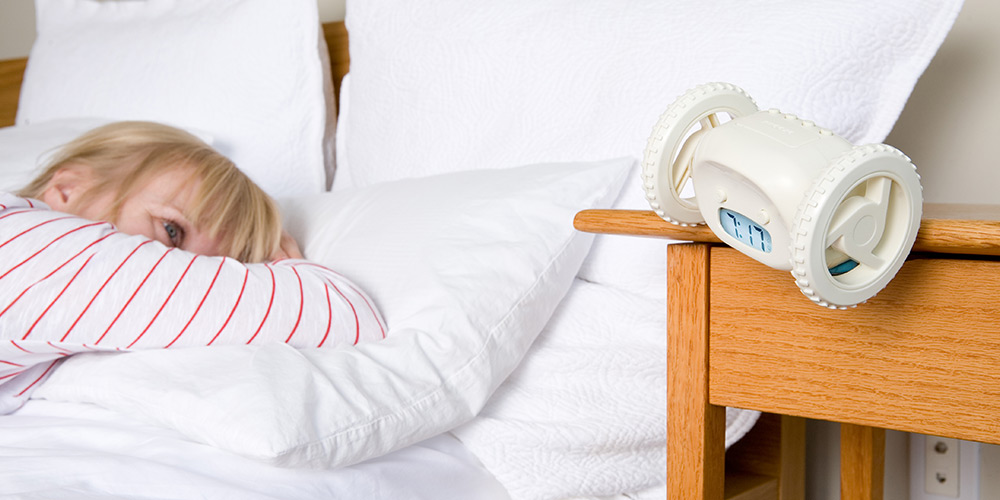 A child in bed looking at the Clocky alarm clock on a wooden bedside table