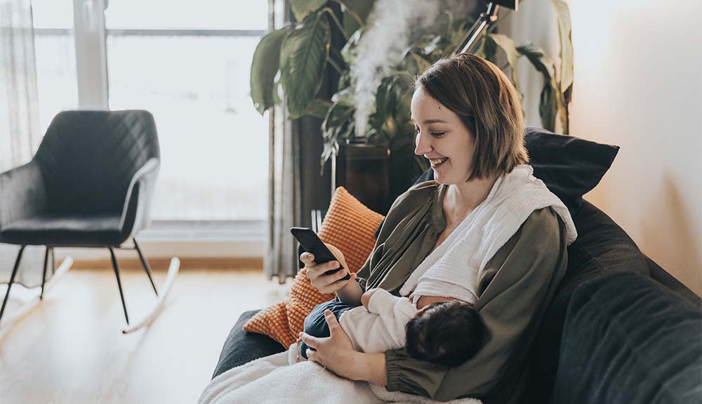 A woman is sitting on a dark coloured couch with a bright orange pillow behind her. She is nursing a baby with dark hair, who is wearing a white shirt and dark pants. She is wearing an olive green shirt with a white towel draped over her shoulder. She is smiling at the cellphone she is holding in her other hand. In the background, steam from a humidifier is visible.