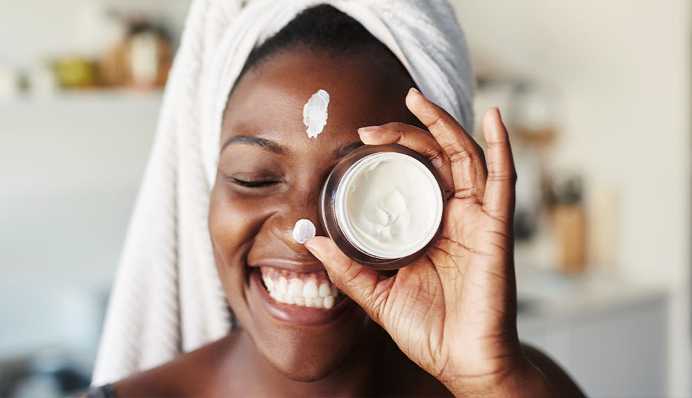 A close up of a woman grinning. She has a white towel wrapped around her head, covering her hair. She is holding up a small dark coloured jar up to one eye. There is a white cream in the jar that is visible. She has a smear of white cream on her forehead above her eyebrows.