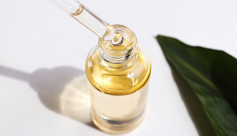 An overhead view of a clear glass bottle with a glass spout. It is filled with a light yellow liquid. Next to it on the white table is a single dark green leaf.