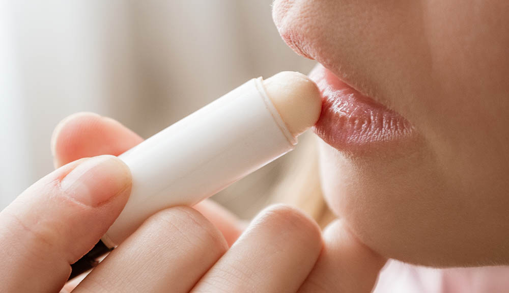 A close up of a person's lips. There is a hand holding up a white tube of lip balm up to the lips. 