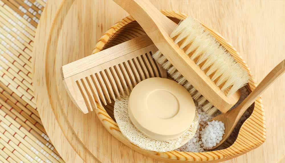 A oval-shaped bowl in a light wood holds several different items in it. There is a larger brush with bristles on both sides in a light wood colour. There is also a smaller brush with bristles with a darker wooden handle. There is a wooden comb and a circular wooden object.