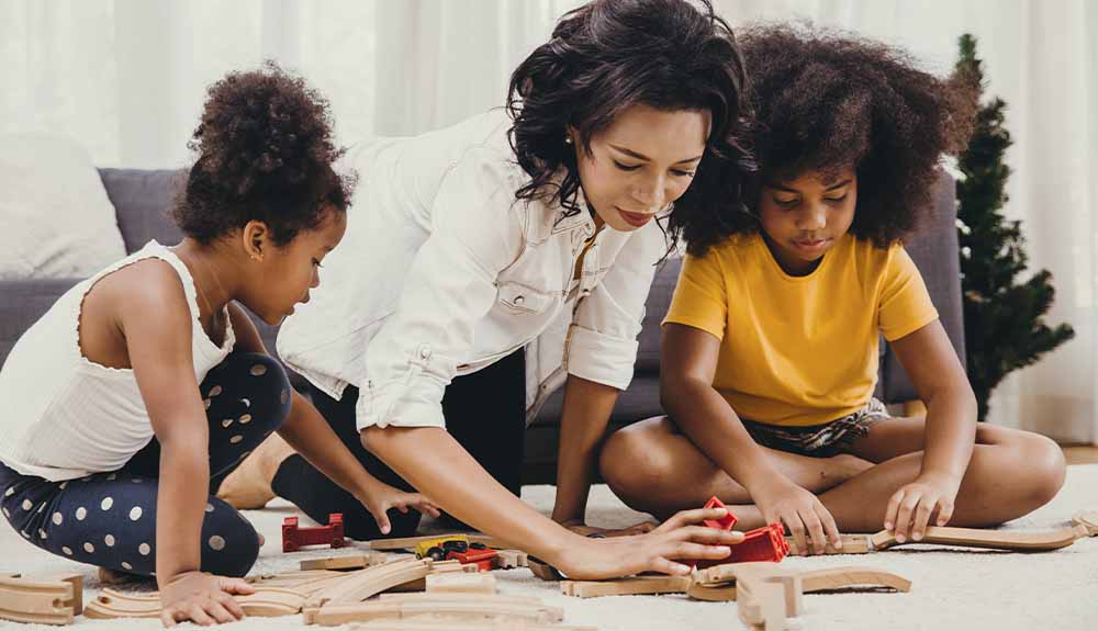 A woman sets up a wooden train track on the floor with two children 