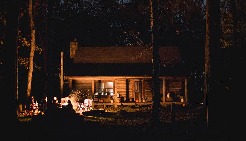 A wood cabin is seen in the glow of a campfire at nighttime, several happy campers sitting around the glowing fire