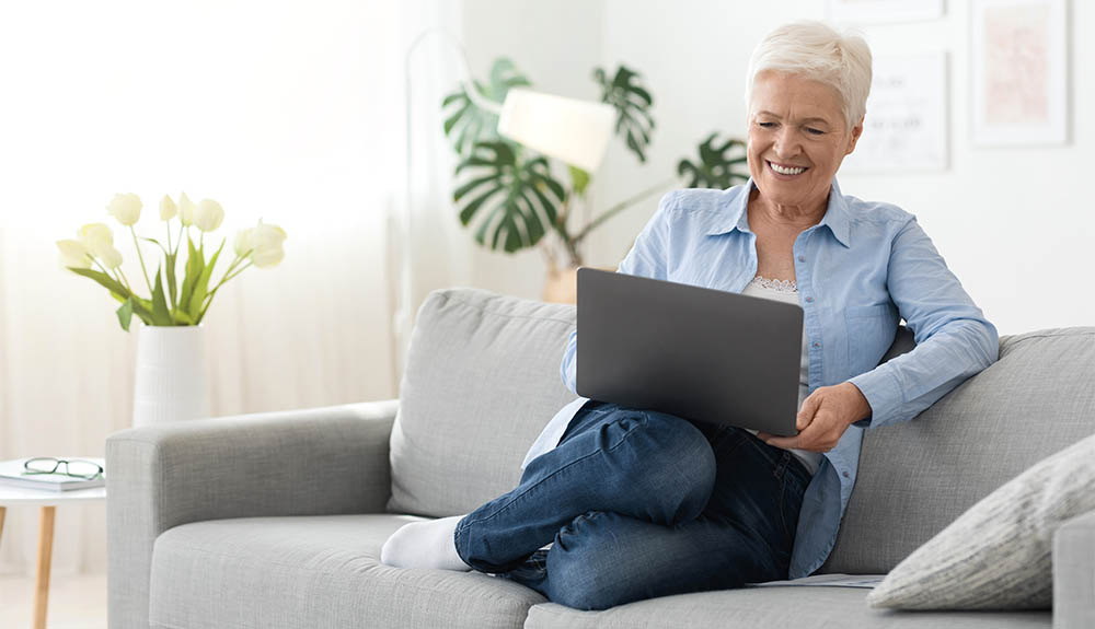 A woman with short white hair wearing a light blue button up shirt that is open with a white top underneath and blue jeans and white socks is sitting on a light grey couch. She is resting a silver laptop on her knees and is smiling at the screen.