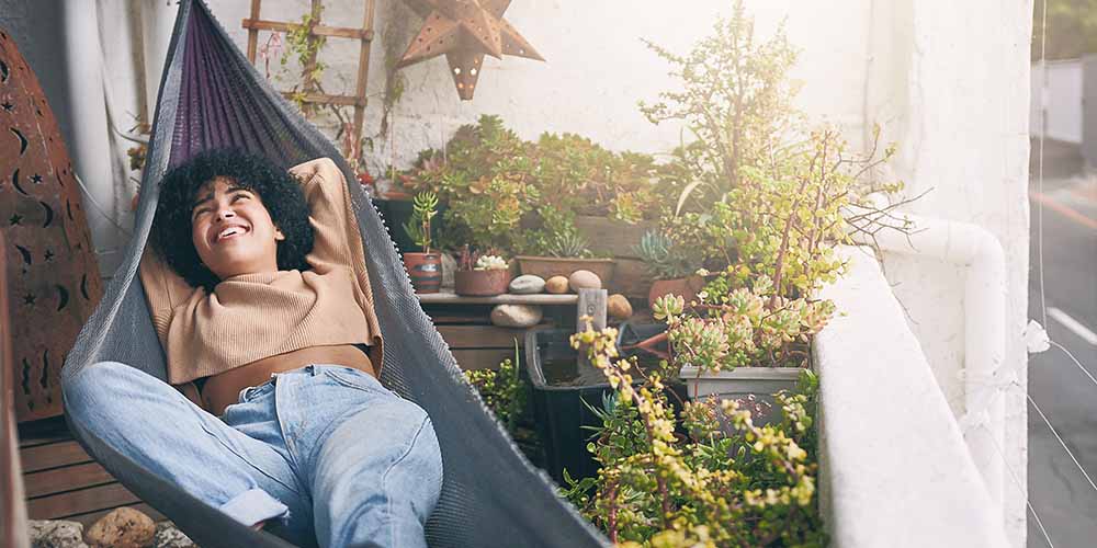 A woman with curly black hair, wearing a peach-coloured crop top and light coloured jeans is lying on a grey hammock with some purple in the corner. She is surrounded by plants on a small balcony, including clay planters with succulents with rocks. 