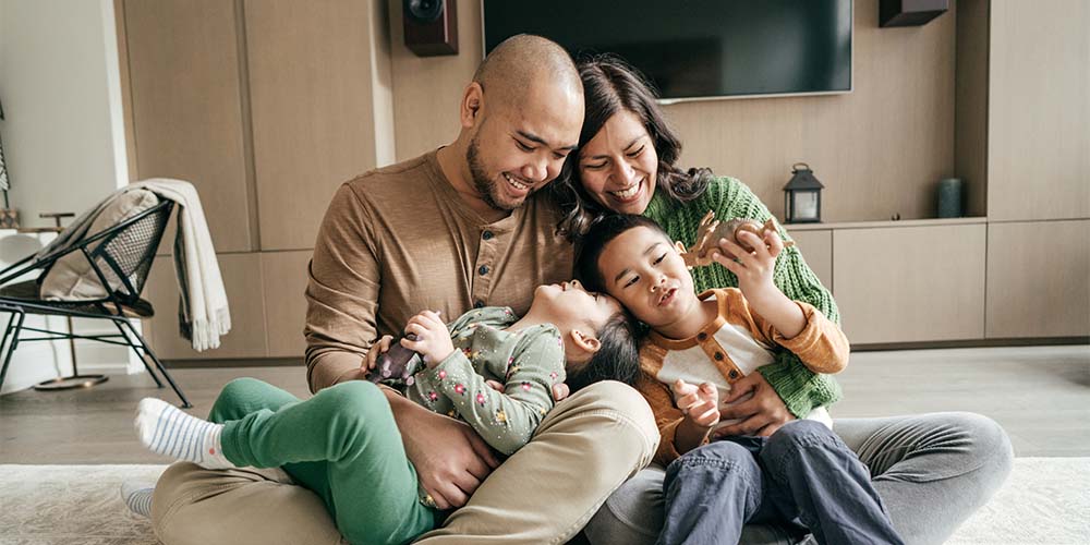 A man in a brown long sleeved shirt and beige pants with a shaved head and small beard and a woman in green knitted green sweater and gray pants with long brown hair sit on a white carpet smiling down at two children. One child is lying on the man’s lap looking upward at him and wearing green pants and a floral long sleeve shirt. The second child is sitting on the woman’s lap, wearing a white and orange log sleeve shirt and jeans and holding a brown dinosaur toy. In the background you see a brown entertainment unit with a TV and a black chair on the left with a pillow and blanket.
