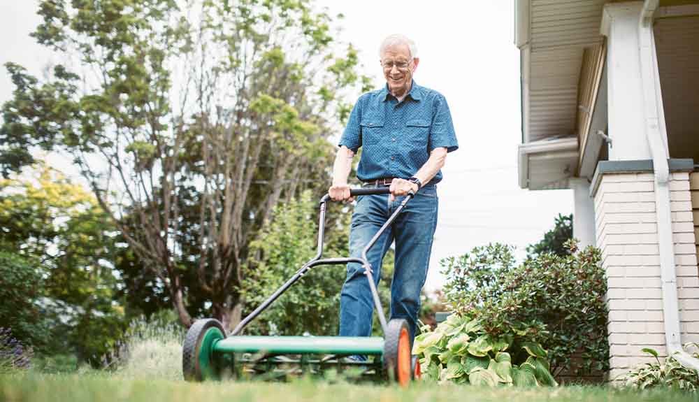 A man with grey hair and glasses wearing a short-sleeved button up blue shirt and jeans with a brown belt. He is holding onto a push lawn mower across some grass. Behind him, a pillar for a home and a porch is visible.