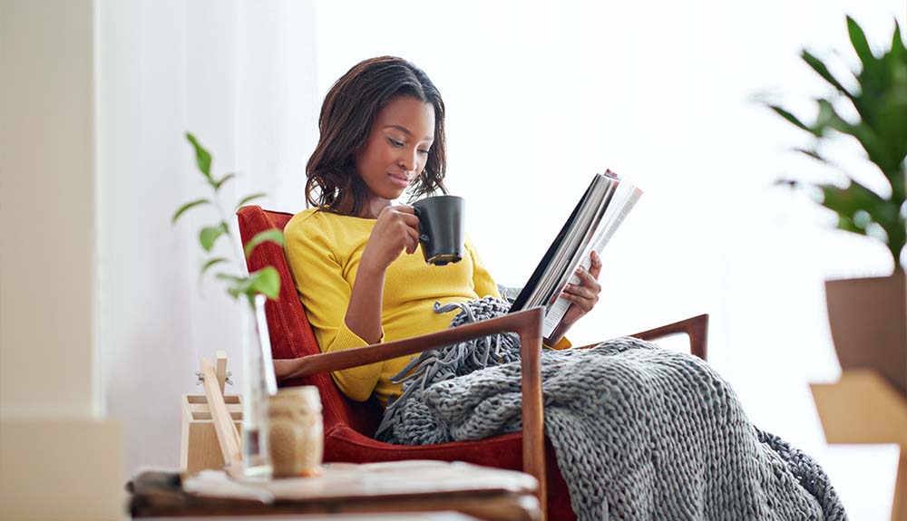 A woman wearing a yellow shirt is sitting in a copper-coloured arm chair. She is holding a black mug and has a grey knit blanket covering her legs. She is looking down at a magazine.
