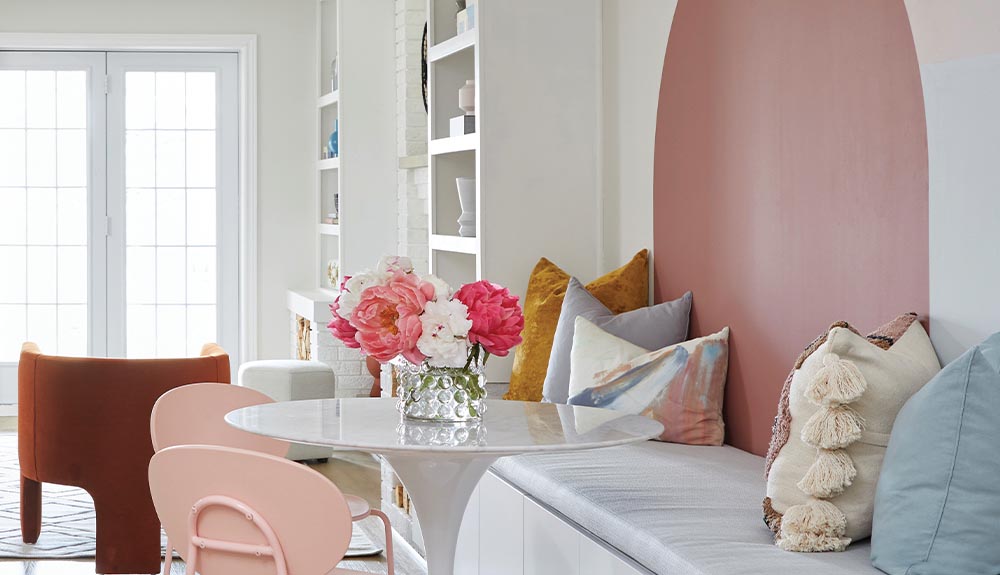 A seating area with a circular white table. There are two blush pink chairs on one side of the table, and on the other is a white banquet with an assortment of decorative pillows. Behind it is a white wall with a painted round pink circle.