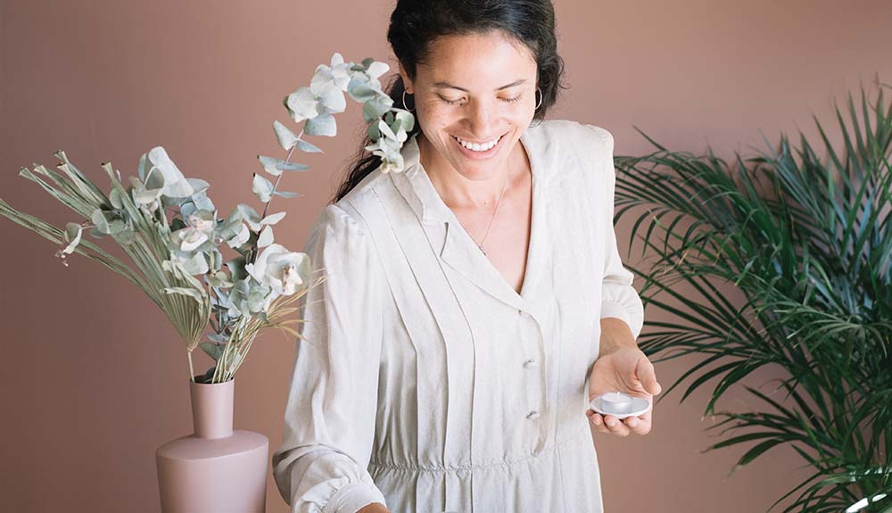 A woman with dark hair wearing a long white button up shirt is standing in front of a pinkish beige wall. There is a beige vase next to her with some leaves and other greenery in it. She is holding a small tea light. There is a larger, leafy plant on the ground behind her.