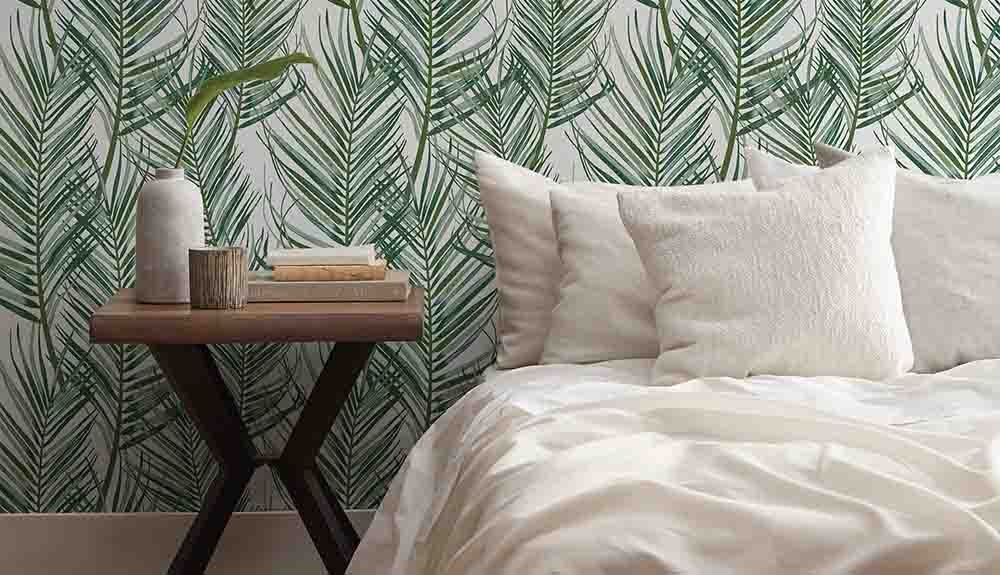 A bed made up with a white comforter and four white pillows with a small table next to it. There are three books on the stand as well as a small white vase with a single leaf in it. Behind the bed is a wallpaper with a leaf motif on it.