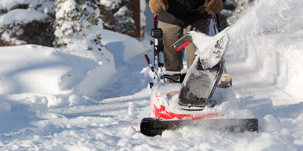 A close up of a red and black snow blower being pushed through some snow. There is a person wearing beige pants and a black coat with thick brown gloves holding onto the handle. There is several inches of snow that has been cleared beside the person, with snow-covered trees.