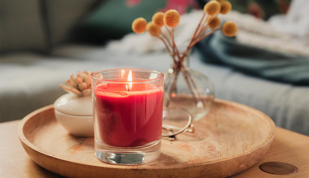 A close up of a circular wooden tray sitting on top of a table. On the tray is a clear glass vase with some yellow floral branches. There is a red candle that's lit and a small decorative object between the vase and the candle. 