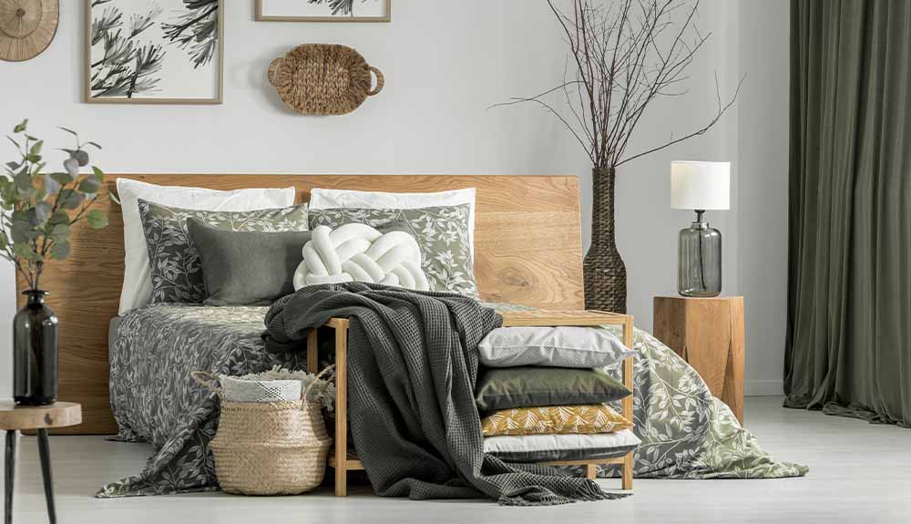 A bedroom with a light wood headboard. There are grey and white sheets, shams and pillows on the bed. On one side of the bed is an oversized dark wicker vase with tall, bare branches. On the other side is a small black vase with leafy green branches sitting on a small wooden side table. Above the bed are two square paintings of branches and wicker decorative baskets hanging.