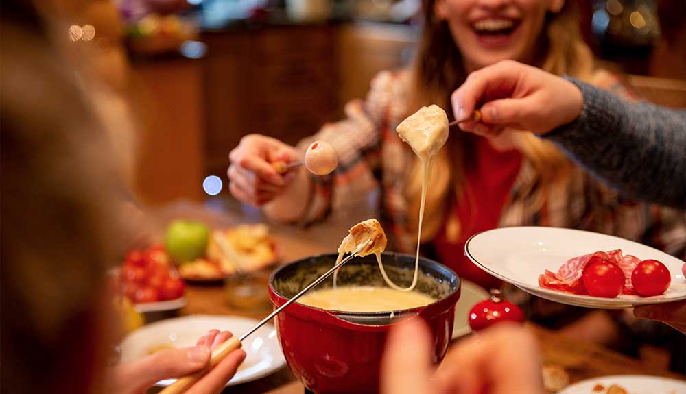 A close up of a red fondue pot with a yellowish cheese in the pot. There is a person with blonde hair wearing a red shirt under an open plaid shirt that is holding something round in her hand. Next to her is a close up of a hand lifting another piece of food out of the fondue pot with a string of cheese attached to it. With the other hand, the person is holding a white plate with some cherry tomatos and cold cuts on it. 