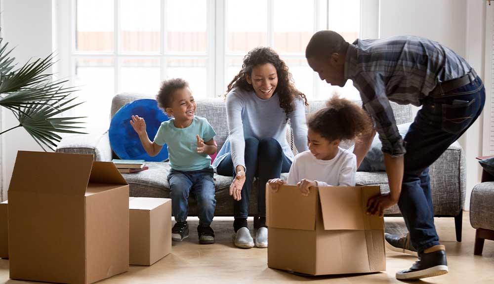 A family of four are pictured in a living room. The dad is pretending to push a little girl wearing a white shirt with a ponytail in a brown cardboard box. A mom and a boy are sitting next to each other on the couch smiling and watching them. There are two boxes of two different sizes next to the boy, who is wearing jeans and a light blue shirt. 