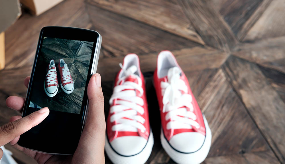 A pair of red converse sneakers on a wooden table, a hand in the corner is seen holding a smartphone, the camera app open and showing a photo of the shoes being taken