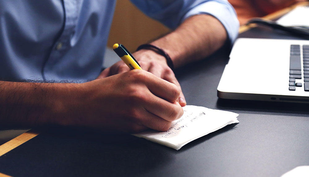 A closeup of hands writing in a notebook, a laptop just to the side