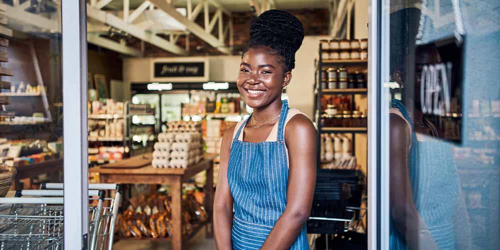 A young woman with black hair in a bun wearing a striped blue and white apron and beige tank top smiles in the doorway of a grocery shop. You can see silver grocery carts on the left, a glass door with an open sign on the right and behind her are tables and shelves stocked with goods. 