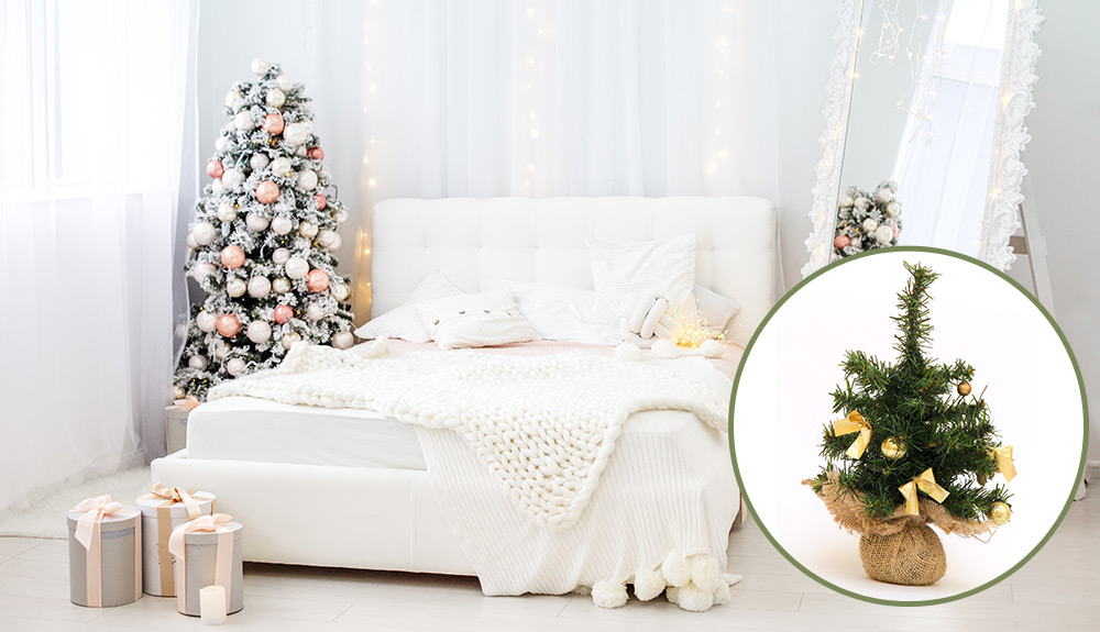 A festive white bedroom with trees at the foot of the bed, twinkle lights behind sheer curtains and a decorated tree in the corner of the room