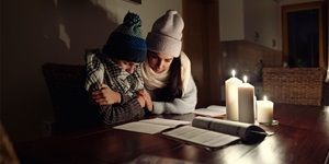 Two people sit at a wooden table with three lit white candles of varying height. They are looking down at white booklets. One wears a striped knitted hat of black, blue and green with a pom-pom on top, a checked black and white scarf and a brown knitted sweater. They have their face in their scarf and arms wrapped around their body. The second sits close with one hand on the other’s arm, and is wearing a white toque, sweater and scarf. 