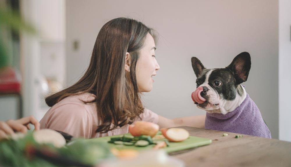 A woman sits at a kitchen table with chopped vegetables on it as her dog sits next to her