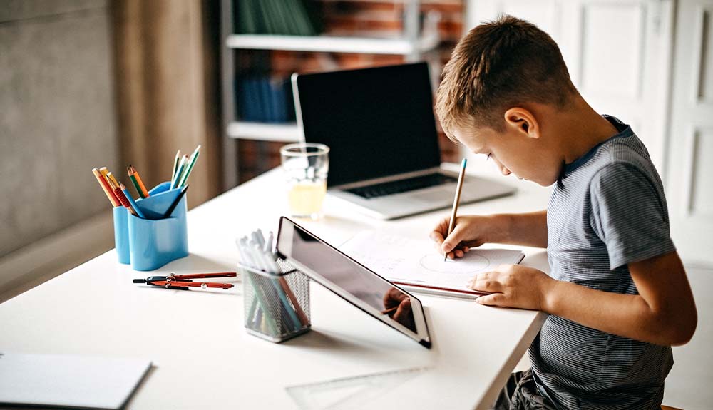 A young boy is writing in a notebook at a desk. Beside him is a tablet on one side and an open laptop on the other side.
