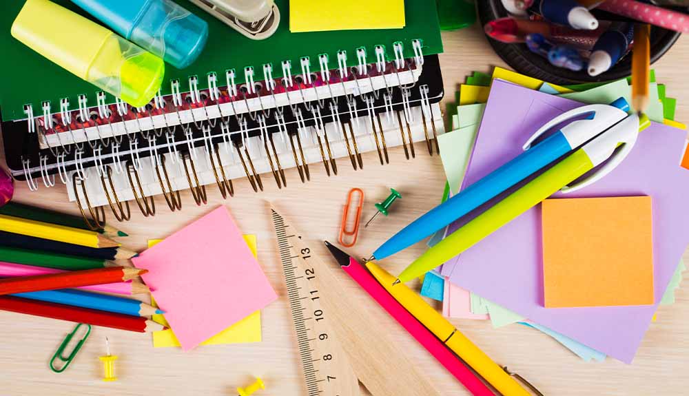 A stack of colourful coiled books on a desk with a blue and yellow highlighter on top of it. Next to it is a stack of purple, yellow and orange sticky notes, with a blue and green pen, as well as a pile of pencil crayons, paper clips and push pins.