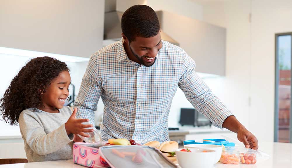 A dad and his daughter standing in a white kitchen. In front of her is a pink lunch bag with rainbows on it. He has a clear plastic container that's filled with grapes and two sandwiches on buns in front of him.