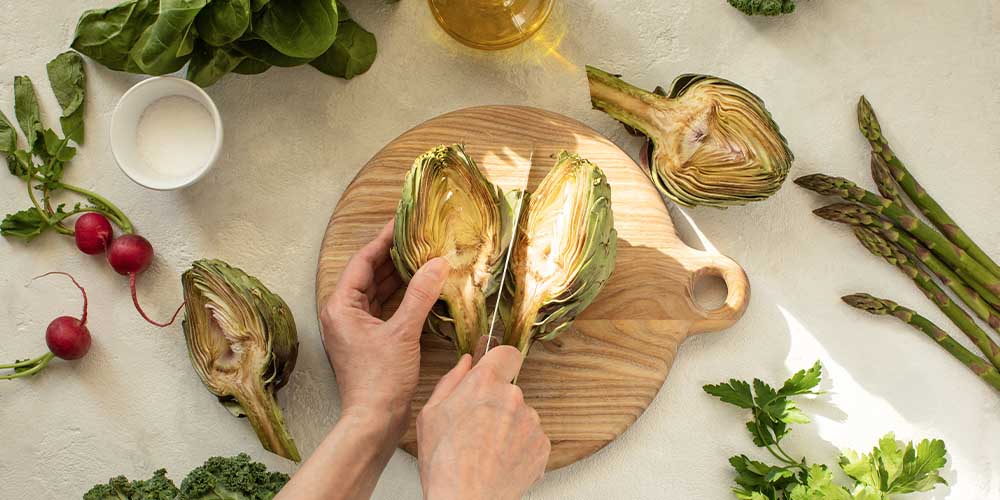 An overhead view of a pair of hands on a counter. There is a circular wooden cutting board with an artichoke cut in half on it. One hand is holding half of the artichoke, with the other hand holding onto a knife and cutting down the middle. Surrounding the cutting board are two halves of another artichoke, as well as some loose asparagus, parsley, radishes and kale. There is a small white container of salt and a bottle of oil in a clear glass container is also visible.