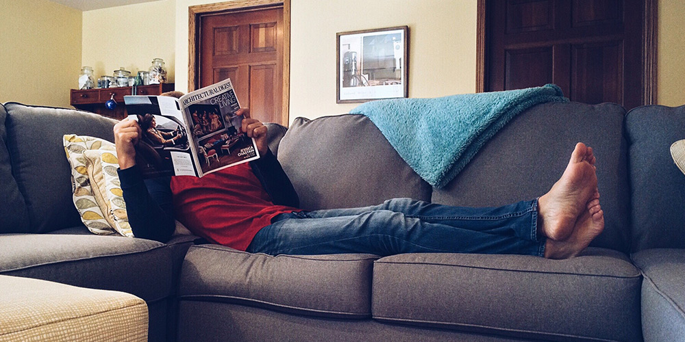 A person lounges on a comfortable grey couch, legs outstretched while reading a magazine
