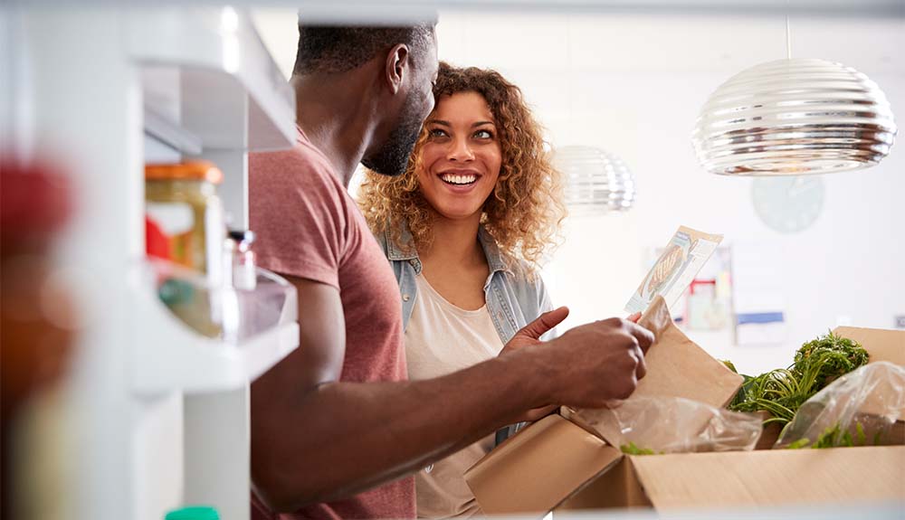 A woman with shoulder-length curly hair is looking at a man who is smiling back at her. They are both standing in front of an opened box with plastic bags and vegetables poking out from the box.