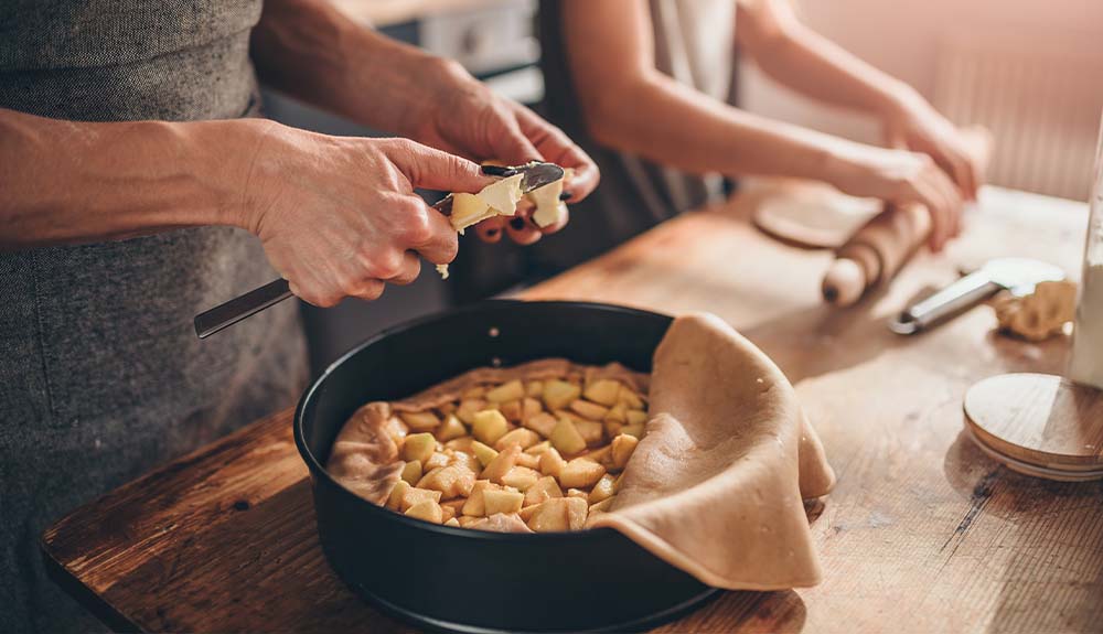 Two people are standing in a kitchen but only their hands are visible. The person in the foreground is cutting bits of butter with a knife into a pie tin. There are chunks of cut apples in the baking tin with some crust hanging over it. The person in the background is rolling out pie crust with a wooden rolling pin.