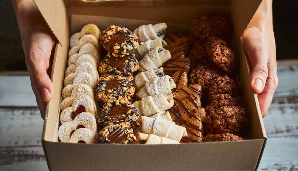 A pair of hands holding a brown paper box of treats. There are four rows of desserts, including heart-shaped cookies with jam and chocolate-drizzled cookies.