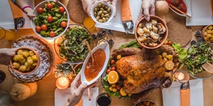 An overhead image of a table laid out with a turkey on a platter with lettuce under it. There are hands holding little bowls of stuffing, sauce, broccoli and tomatoes and olives alongside bowls of salad. 