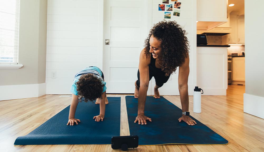 A woman with curly dark hair is with a toddler wearing a striped turquoise and navy T-shirt. They are both on dark teal yoga mats on the floor, in the plank position. The mom is looking at her child and smiling. There is a white water bottle next to the mom, and a black cell phone in front of them.