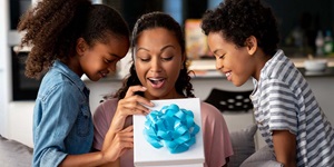 A woman is sitting between two school-aged kids. She is looking down at a box that is wrapped in white paper with a large blue bow on the lid. She is smiling in surprise, and the kids are looking inside and smiling as well.