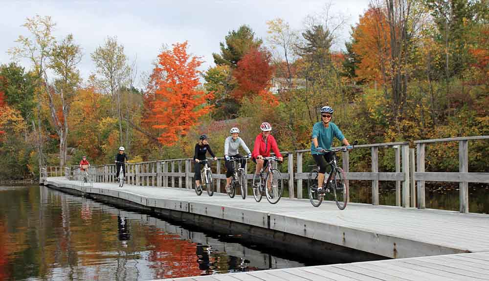 A row of cyclists, one behind the other, are riding their bikes wearing helmets on a boardwalk next to some water. Behind them, the trees are changing colours into shades of orange and yellow.