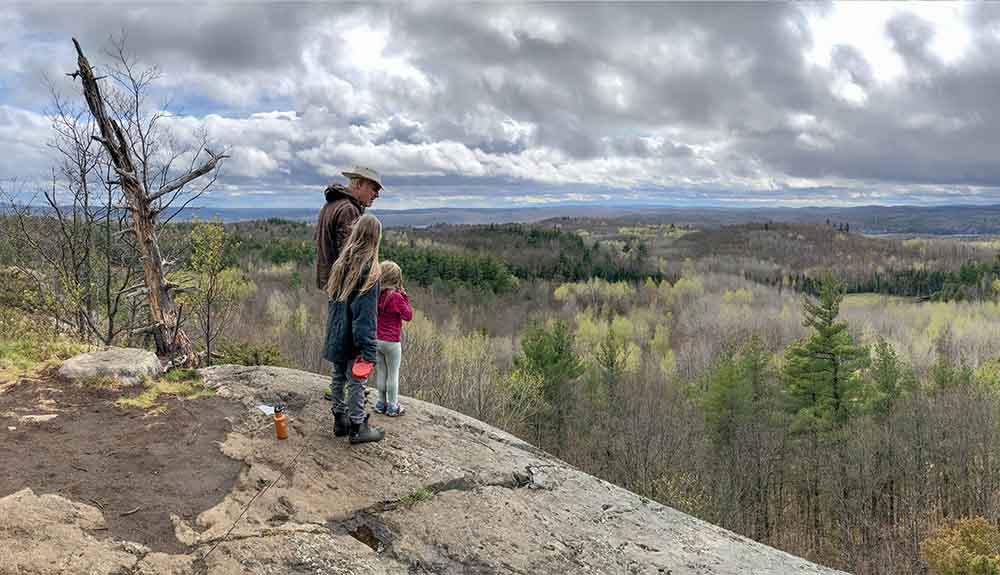 An older man wearing a khaki hat and a brown coat with a hood is standing at the edge of a rock with two young kids next to him. There is a little girl in front wearing a dark pink hoodie and light green leggings. There is a taller girl behind her wearing a long blue coat and grey pangs. They are all looking down at the trees below them.