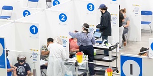 A row of partial cubicles with white numbers in blue circles marking 1, 2 and 3. There are two people in each of the cubicles. One person is sitting down while another person is standing next to them. Some are leaning over the other person. There are carts of medical supplies next to each one person.