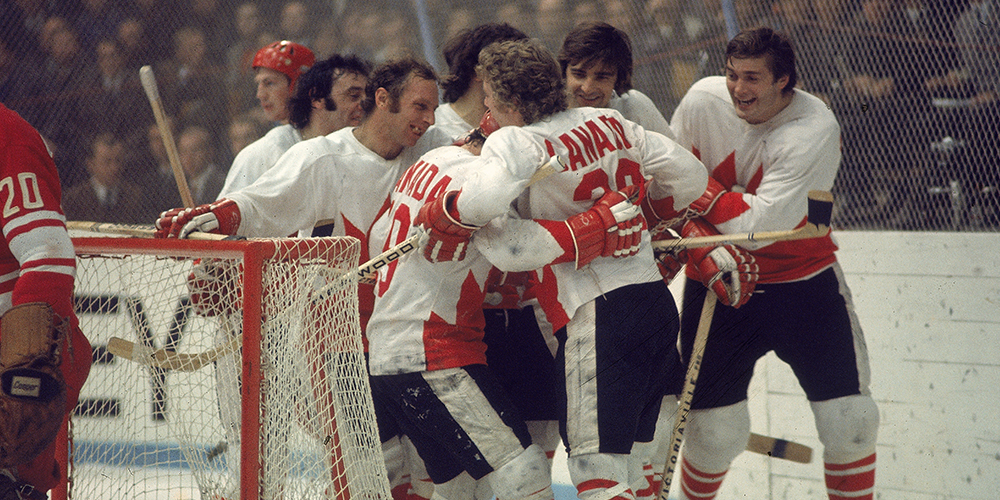 Canadian hockey players engage in a group hug wearing white and red uniforms after a game behind the net