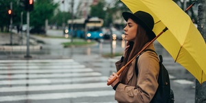 A woman with long brown hair wearing a beige coat and a black backpack holding a bright yellow umbrella. She is standing at a crosswalk that looks wet. There is a blue city bus behind her stopped at an intersection.