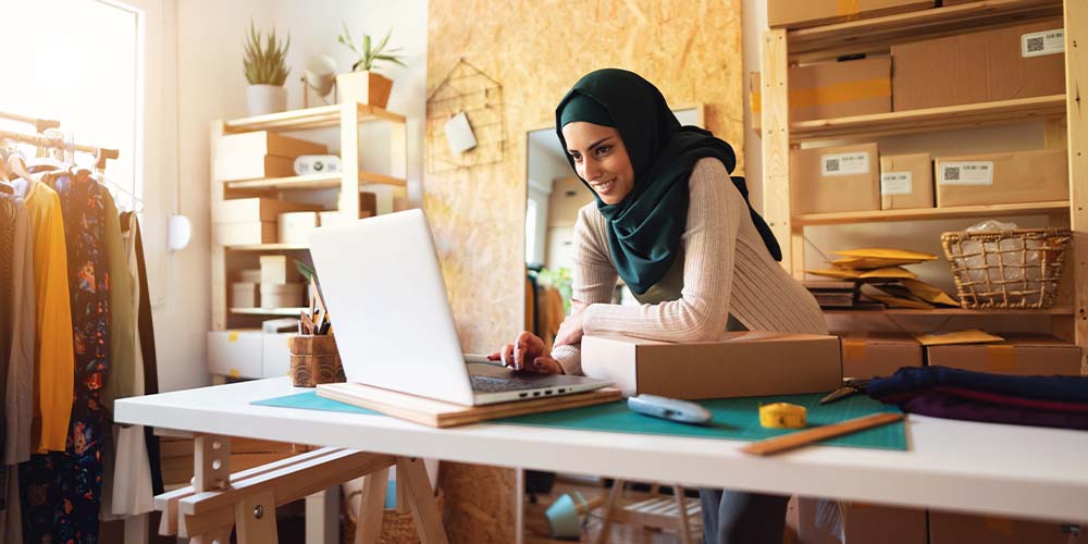 A woman wearing a dark green hijab, light pink cardigan and green shirt is standing in front of a laptop on a workbench-style table. She is looking at a laptop and smiling at the screen. One arm is positioned over a cardboard box. Behind her are two wooden shelves. There is one in a corner by a window filled with boxes of different shapes and sizes. Some of the boxes on the shelves directly behind her have mailing labels on them. There is also a full-length mirror leaning on a wooden wall behind her and a rack of clothes.