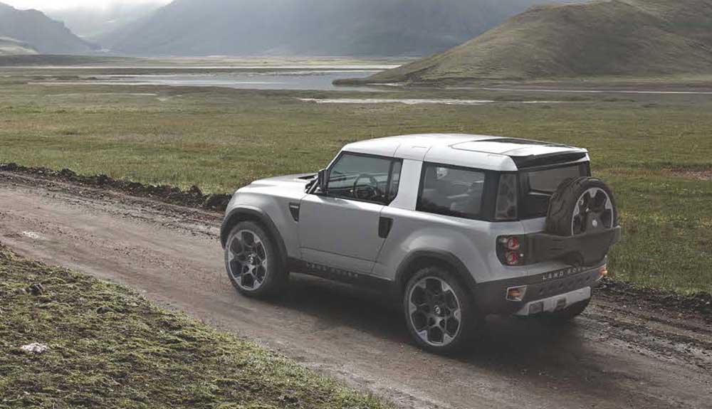 A silver Land Rover Defender is shown on a country road with hills in the background