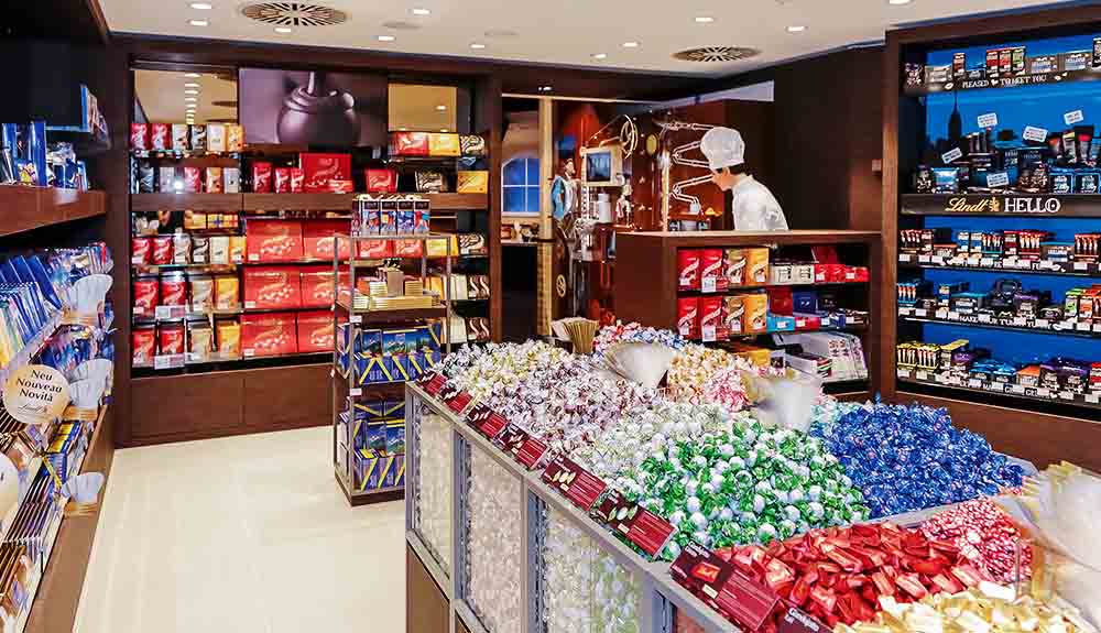 The interior of the Lindt chocolate shop in Jungfraujoch, Switzerland, plastic display cases piled high with colourful chocolates and a man in chef whites seen behind a counter