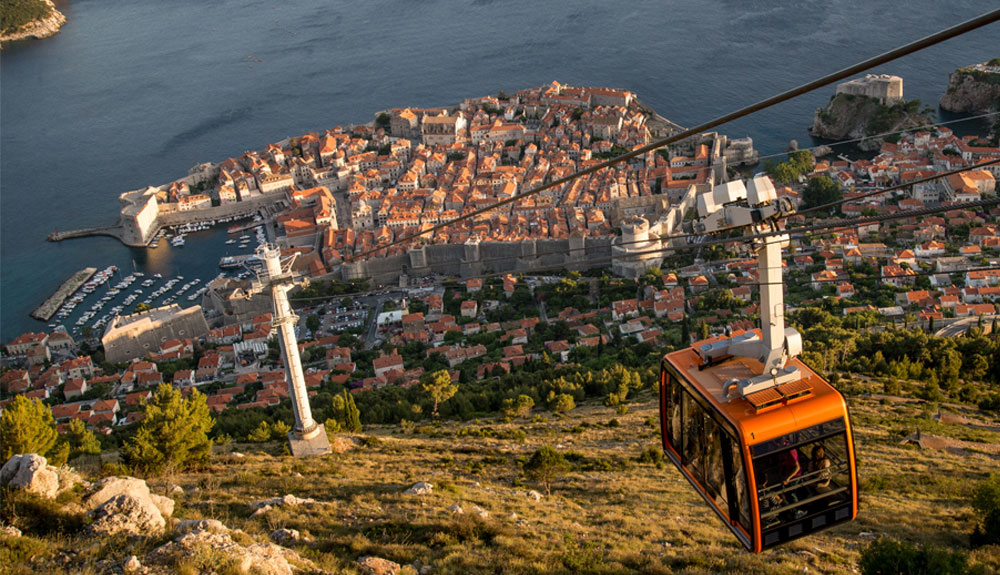 A gondola is shown making its way down a mountainside with the ocean in the background
