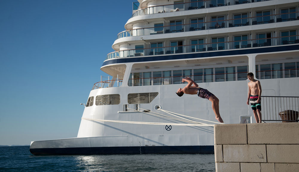 Man dives backwards off a dock in front of a large cruise ship in Zadar, Croatia