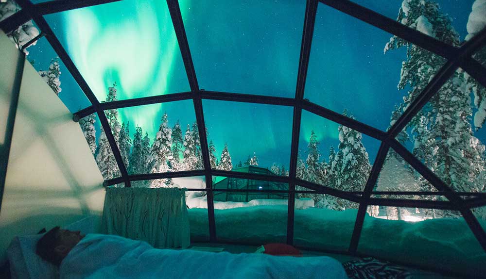 A person snuggles in bed in a heated glass Igloo at Kakslauttanen Arctic Resort, the green glow of the aurora borealis and snowy trees visible through the glass