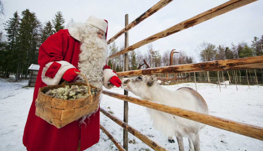 A man dressed as Santa feeds a reindeer behind a wooden fence at Santa's Village in Finland